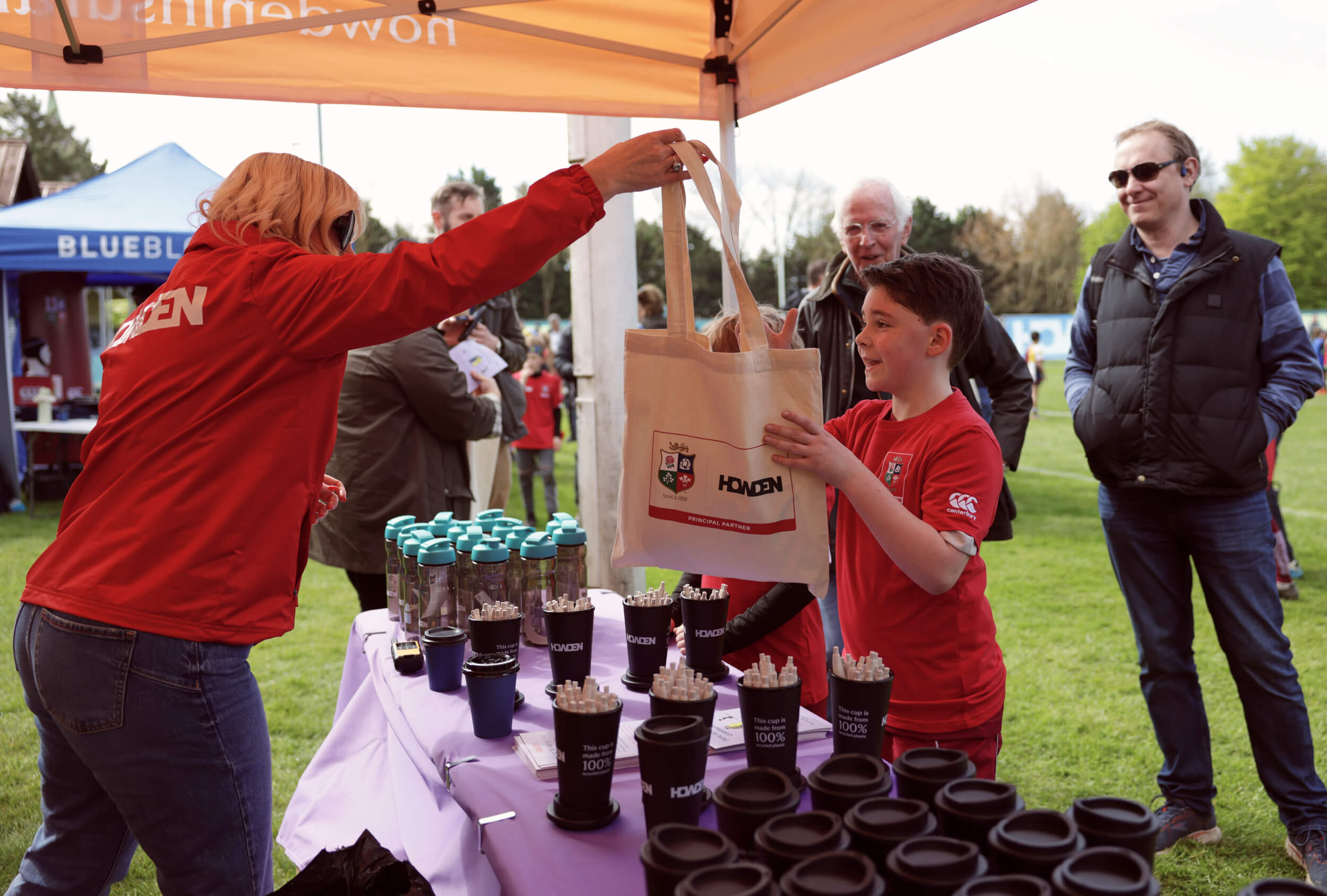 A woman hands a tote bag to a young boy at a booth displaying reusable Howden branded cups during an outdoor event. Several people stand nearby.